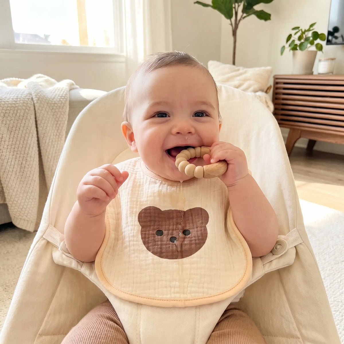 bébé dans un transat avec un anneau de dentition dans la bouche et un bavoir en gaze de coton motif ourson sur lui