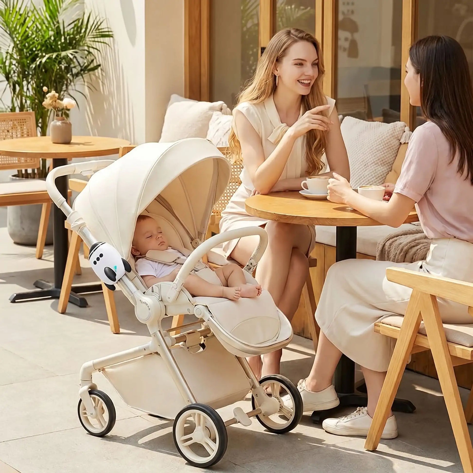 deux femmes qui boivent un café en terrasse avec un bébé qui dort dans une poussette et un berceur électrique pour poussette avec connexion Bluetooth fixé dessus