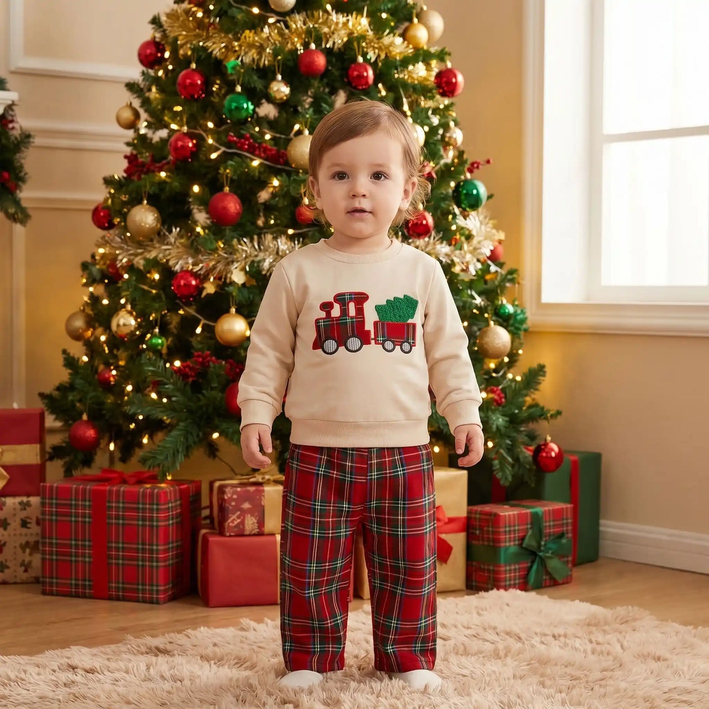 petit garçon debout dans un ensemble de noël bebe garçon en coton avec un pantalon à carreaux rouge et un sweat avec un petit train qui tire un sapin