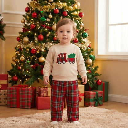petit garçon debout dans un ensemble de noël bebe garçon en coton avec un pantalon à carreaux rouge et un sweat avec un petit train qui tire un sapin