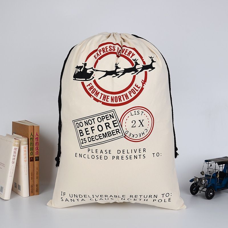 Une hotte du Père Noël réutilisable en toile de coton avec un cordon de serrage noir. L'impression présente un sceau circulaire central avec un traîneau et des rennes, entouré du texte "EXPRESS DELIVERY FROM THE NORTH POLE". Tampons "DO NOT OPEN BEFORE 25 DECEMBER" et "LIST 2X CHECKED". Une ligne est prévue pour le nom sous "PLEASE DELIVER ENCLOSED PRESENTS TO:". Le sac est rempli et posé sur une surface blanche