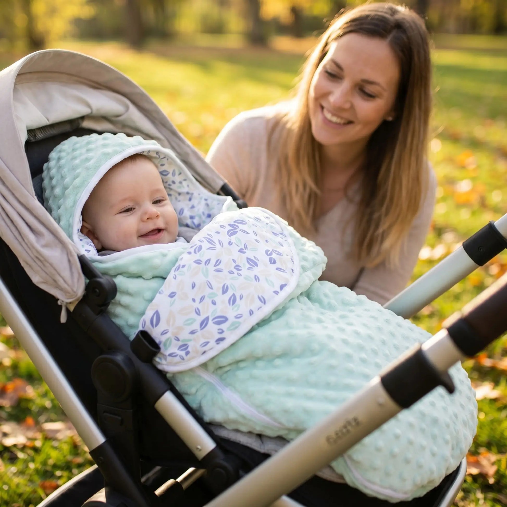 maman souriante au parc qui regarde son bébé dans sa poussette attaché enveloppé dans son nid d'ange cosy vert avec des feuilles imprimées