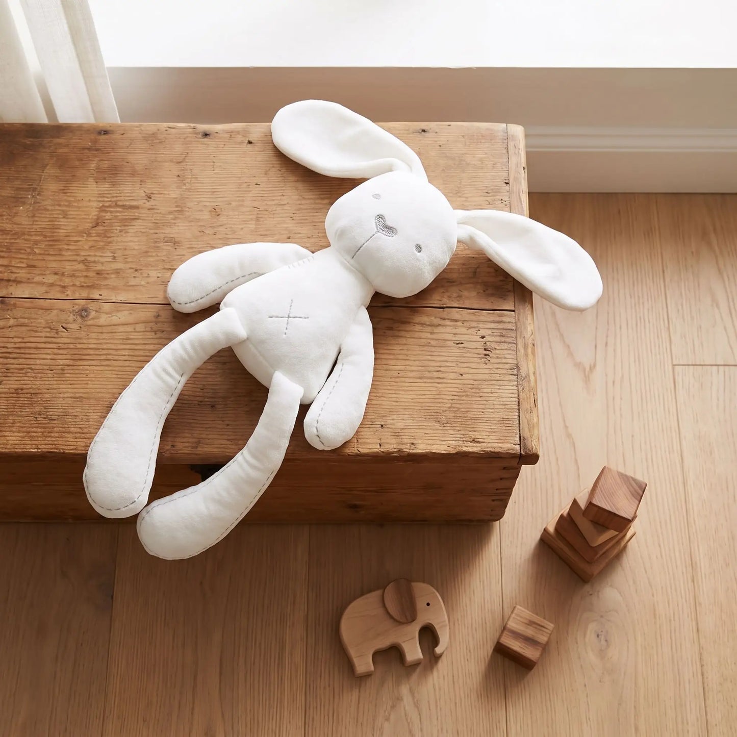 Doudou lapin blanc de naissance posé sur un meuble en bois, montrant ses détails brodés sécuritaires et ses coutures renforcées.