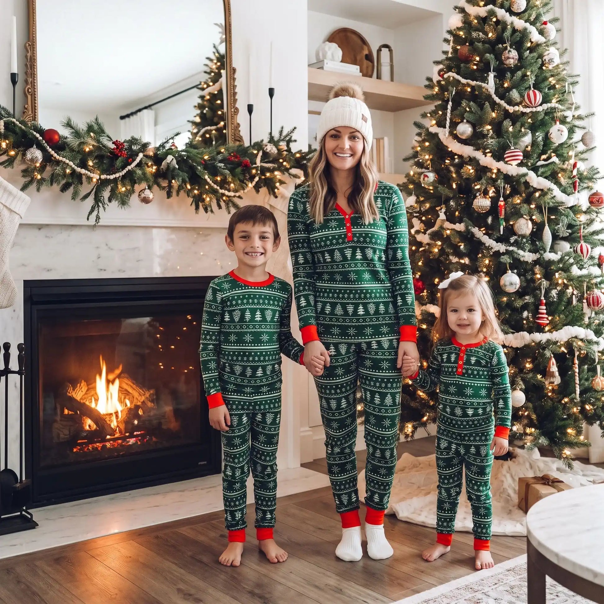 une maman souriante tient les mains de chaque côté de ses enfants tous habillées en pyjama de noel assorti famille sapin vert 