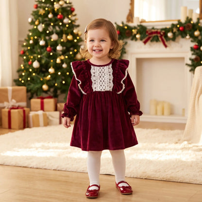 Robe de Noël bébé fille en velours rouge bordeaux avec plastron en dentelle blanche et volants, portée par une petite fille souriante avec des collants blancs et des petites ballerines rouges par la boutique Petitpas et maman