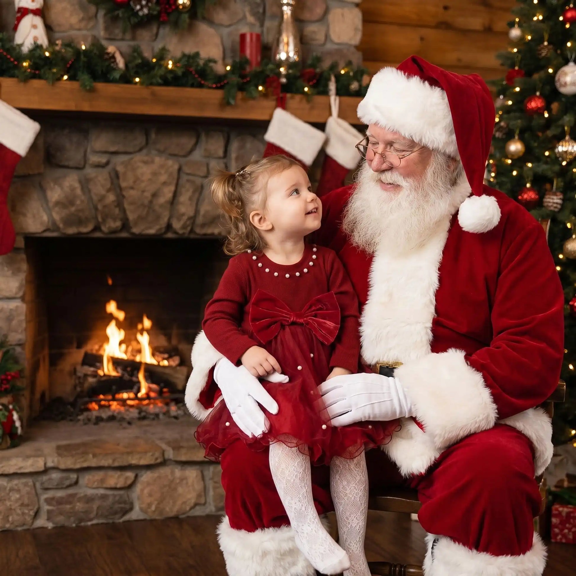 petite fille sur les genoux du père noël habillée d'une robe de noël bébé fille et des collants en dentelle blanc avec en arrière plan une cheminée en pierre et un feu allumée