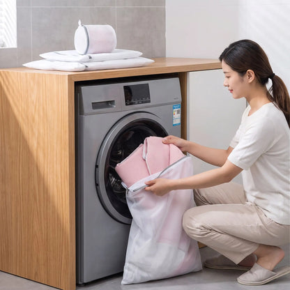 maman accroupit devant sa machine à laver frontale grise qui met dans un sac de lavage grand format un tour de lit rose