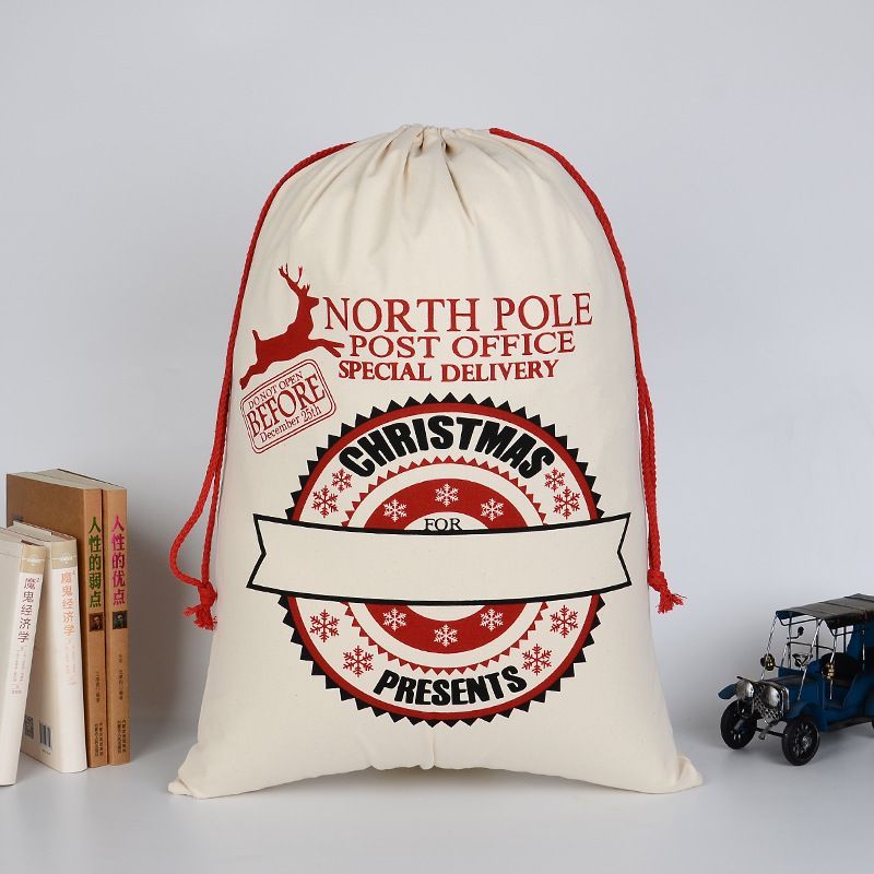 Un grand sac de Noël en coton écru réutilisable, avec un cordon de serrage rouge épais. Le design vintage imite un colis postal avec un grand sceau "NORTH POLE POST OFFICE SPECIAL DELIVERY" et un ruban "CHRISTMAS" personalisable. Tampon "DO NOT OPEN BEFORE December 25th" avec motif élan. Le sac est rempli et posé sur une surface blanche à côté de livres