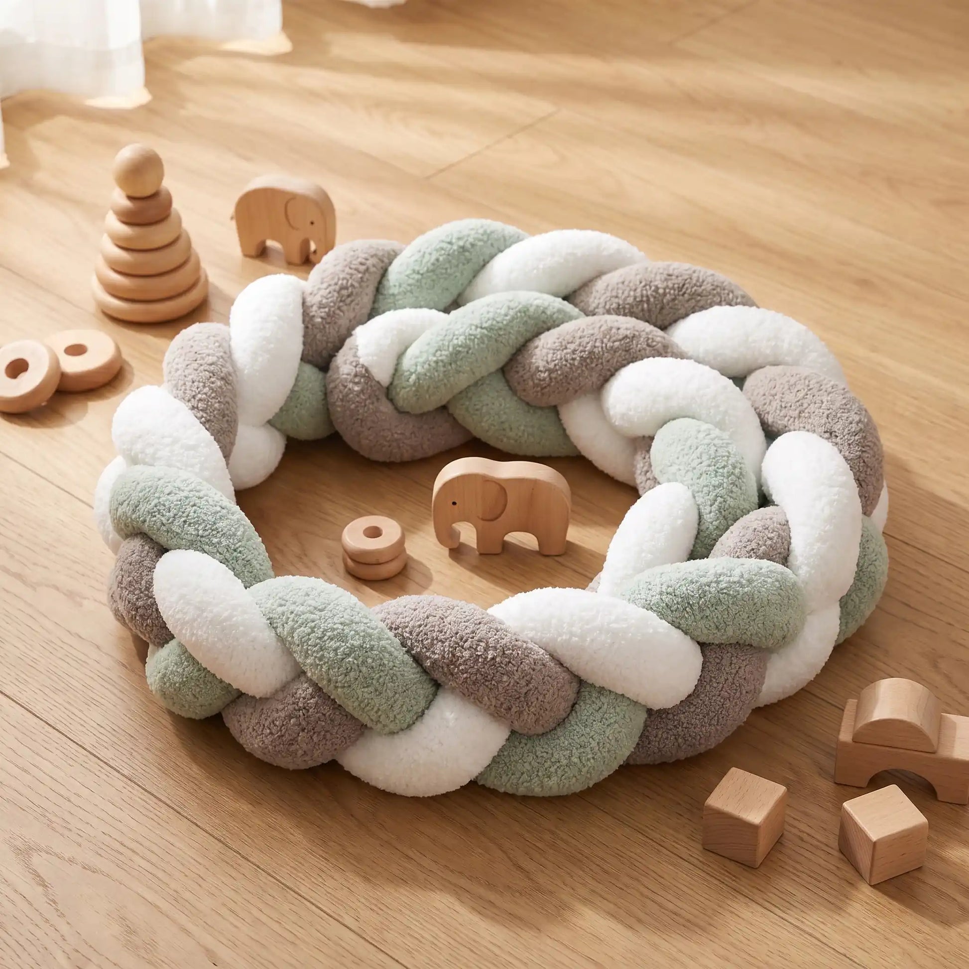 Tour de lit tressé vert sauge pour bébé, enroulé sur un parquet en bois à côté de petits jouets d'éveil en bois.