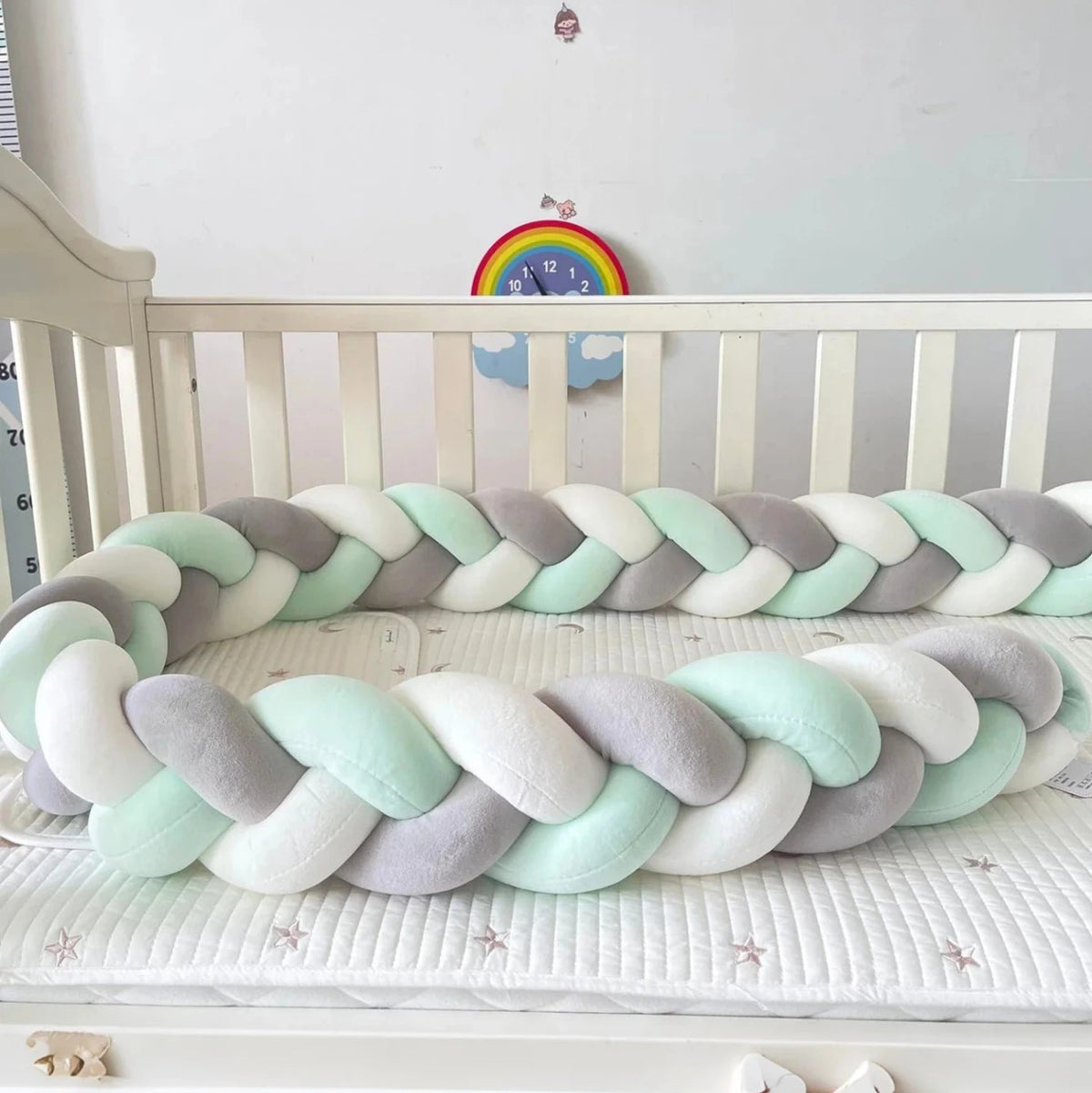 un tour de lit tressé vert sauge gris et blanc de 2 mètres posé contre les barreaux d'un lit bébé à barreaux blanc