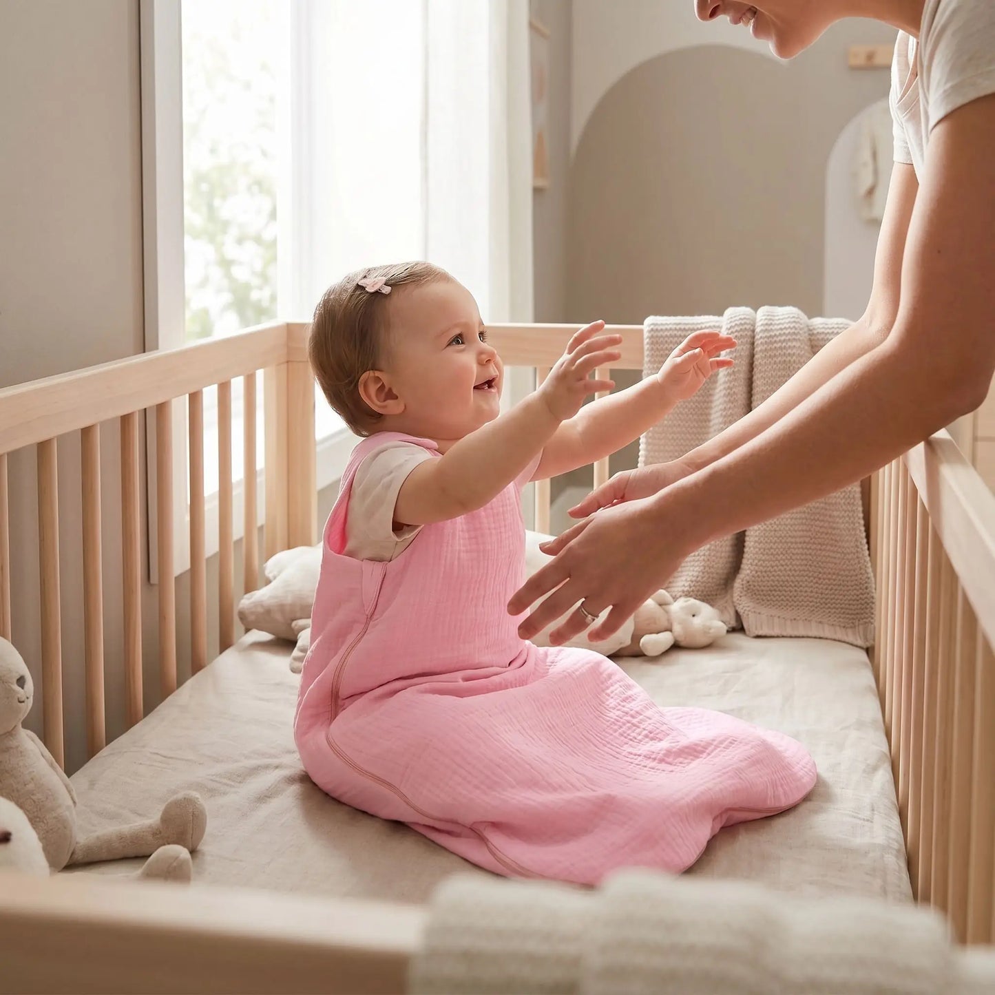 bébé assis dans son lit habillé d'une turbulette été pour une chambre de plus de 21 degrés qui tend les bras vers sa maman 
