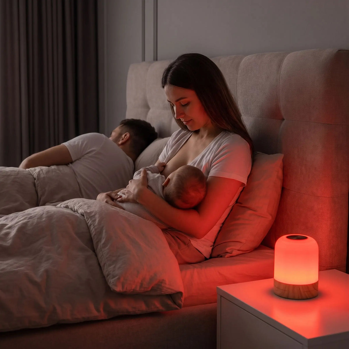 Une maman allaite son bébé au sein pendant la nuit, assise dans le lit conjugal. Sur la table de chevet, une veilleuse bébé lumière rouge diffuse un éclairage doux et sans lumière bleue, permettant à la mère de voir clair sans réveiller le bébé ni son partenaire qui continue de dormir paisiblement à côté d'elle.