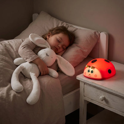 petite fille qui dort paisiblement avec son doudou lapin longues oreilles 45 cm dans ses bras  avec une veilleuse bébé lumière rouge coccinelle posée sur une table de chevet qui émet une lumière rouge tamisée 