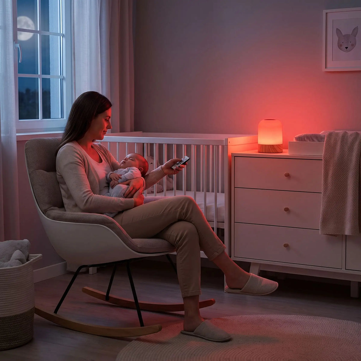 Une maman assise sur un fauteuil à bascule dans la chambre, berçant son bébé endormi de nuit grâce à une veilleuse bébé lumière rouge 670 nm posée sur une commode près du berceau. Elle utilise la télécommande pour régler l'intensité de la douce lueur rouge apaisante, idéale pour les réveils et tétées nocturnes sans perturber le sommeil.