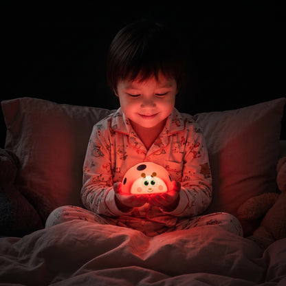 petit garçon assis dans son lit qui sourit et regarde sa veilleuse coccinelle lumière rouge apaisante favorisant la production de mélatonine