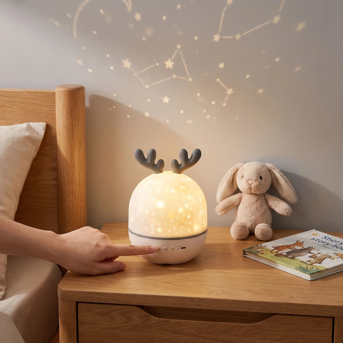 veilleuse musicale bébé projecteur ciel étoilé posé sur une table de chevet de format compact à côté d'un doudou et d'un livre d'enfant